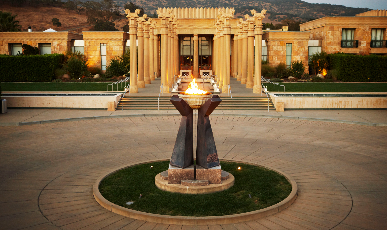 Exterior of winery with large sculpture in center of circle drive holding a basin with lit fire in front an entrance lined with columns