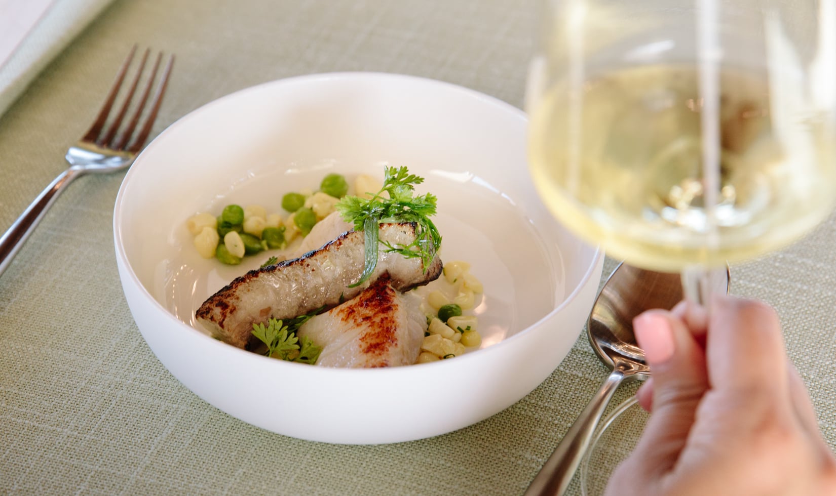 How to Read a Wine List Image close up of halibut dish with a hand holding a glass of chardonnay wine