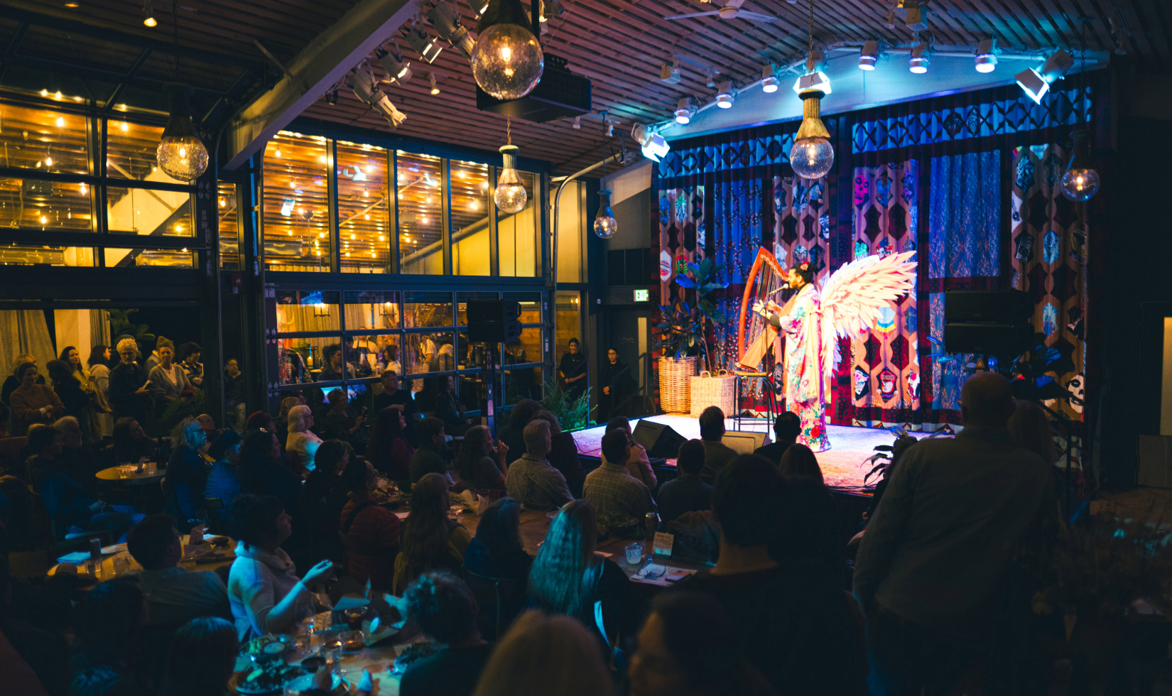 Audience watches performance on a stage in an intimate venue where a performer, dressed in a colorful robe and pink wings, plays the harp and sings
